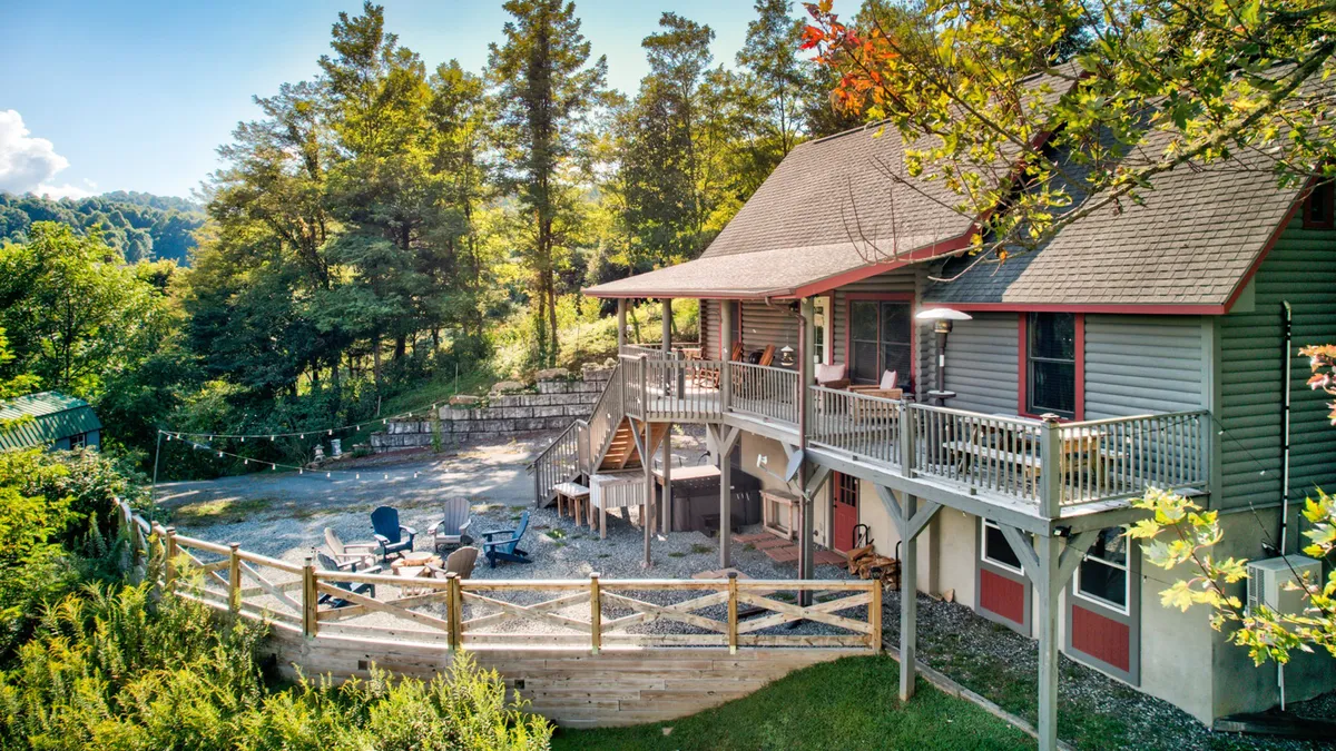 A welcoming exterior shot of the Great Bear Lodge, featuring the house nestled among tall trees. The spacious deck is visible with comfortable outdoor seating, ideal for owners and pets alike. The surrounding natural landscape hints at nearby trails and pet-friendly outdoor adventures.