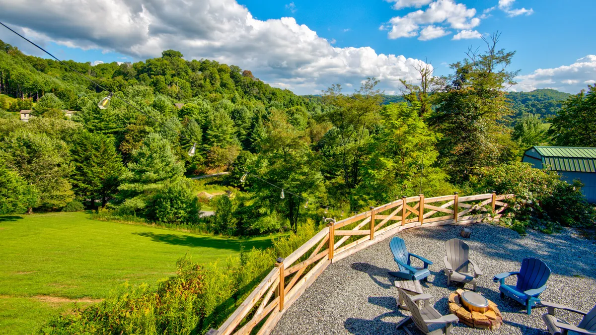 An exterior view of the fire pit area surrounded by Adirondack chairs, set on a gravel patio with lush greenery and a stunning backdrop of mountain views. The setting sun casts a warm glow, ideal for evening gatherings and fireside stories.