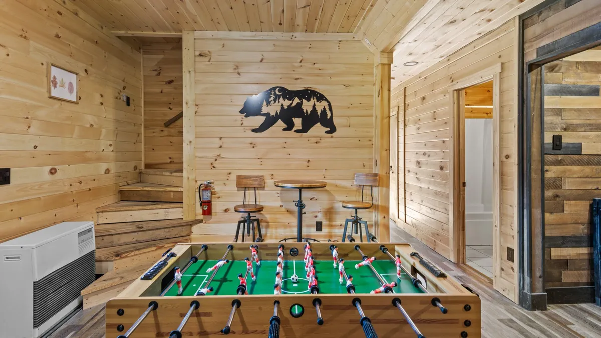 This interior image captures the lively recreation room with a foosball table as the centerpiece, surrounded by wooden bar stools and a distinctive bear silhouette wall feature. People are shown playing foosball, socializing, and relaxing, setting a playful and welcoming tone.