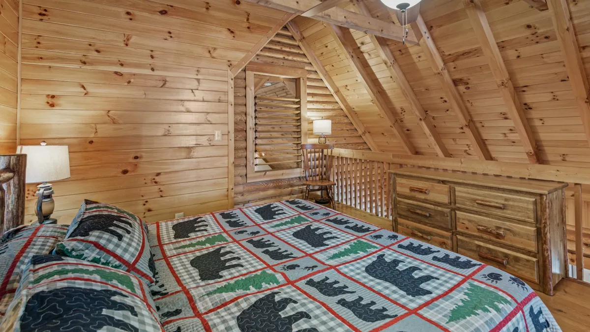 A bedroom with a wooden ceiling, bear-patterned quilt on the bed, wooden dresser, and a soft lamp. The space feels tranquil and private, with rustic decor that suits a mountain getaway loft suite.