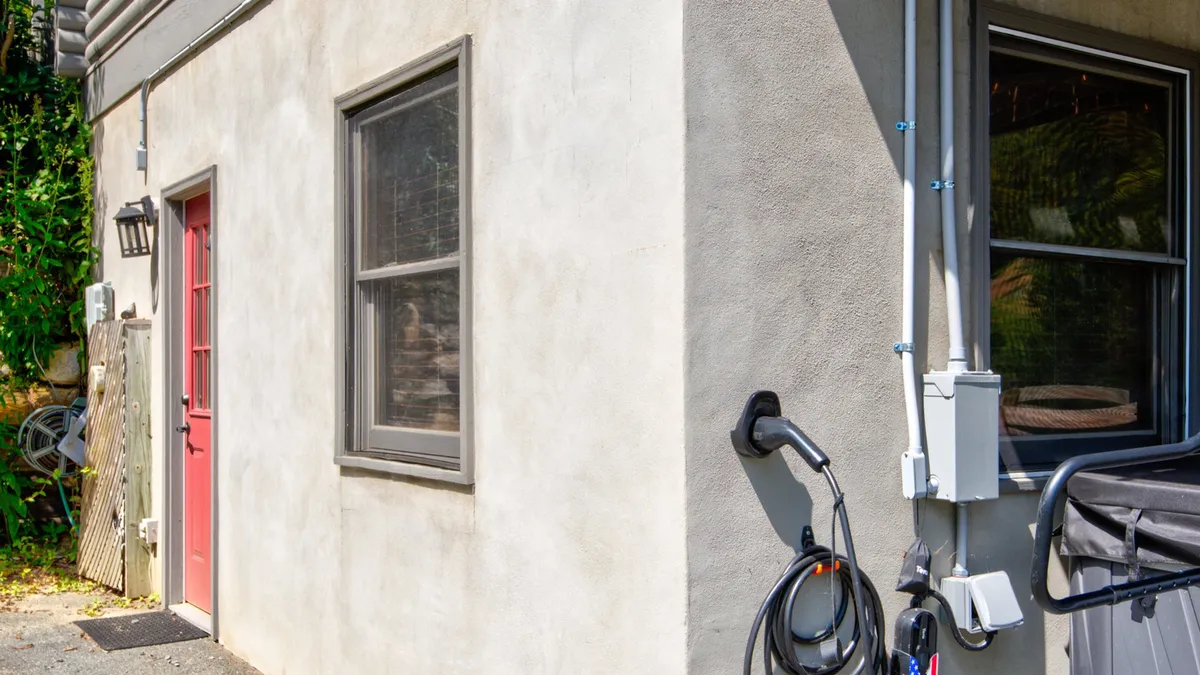 The photo captures the home's entryway, featuring a red door, windows, and a visible electric vehicle charging station adjacent to the paved parking area. The mood is welcoming and hints at eco-friendly conveniences.