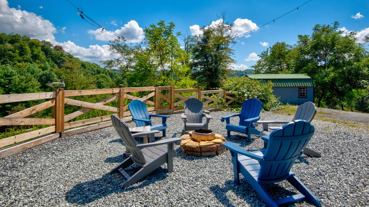 A cozy fire pit area is surrounded by adirondack chairs and a rustic wooden fence, nestled among tall trees. The space encourages guests to gather, share stories, and enjoy evening s'mores or stargazing sessions under the open sky.