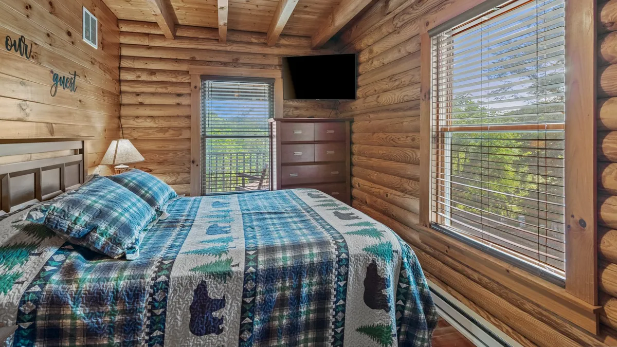 A cozy bedroom featuring a bed with a patterned quilt, wooden walls, a window with blinds letting in natural light, and a wall-mounted TV. The serene and comfortable atmosphere is enhanced by the use of warm wood tones throughout the space.