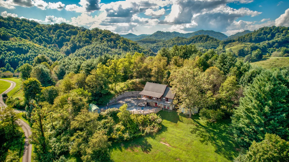 The exterior of the lodge sits peacefully among towering trees with a sweeping mountain backdrop and ample open space. The porch offers sunset views, and the landscape suggests both tranquility and easy access to hiking and nature exploration.