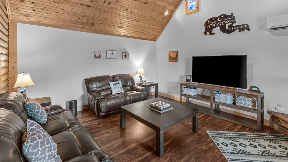 This image showcases a living room with plush leather sofas arranged around a wooden coffee table, facing a large TV unit. Decorative wall art and a cozy lamp add a welcoming touch, creating an inviting atmosphere perfect for movie nights or family gatherings.