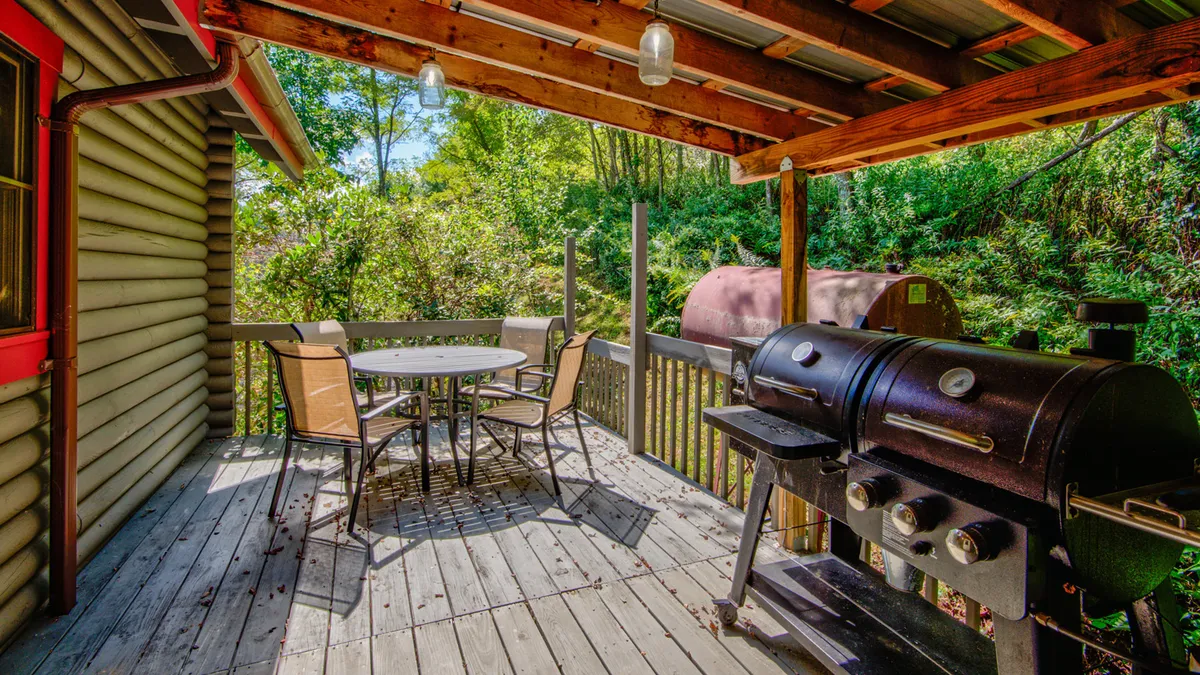 A grill is set up on the deck next to a dining table, surrounded by greenery. The setup is casual yet inviting, ideal for barbecues, relaxed meals, and enjoying nature's beauty just steps from the interior.