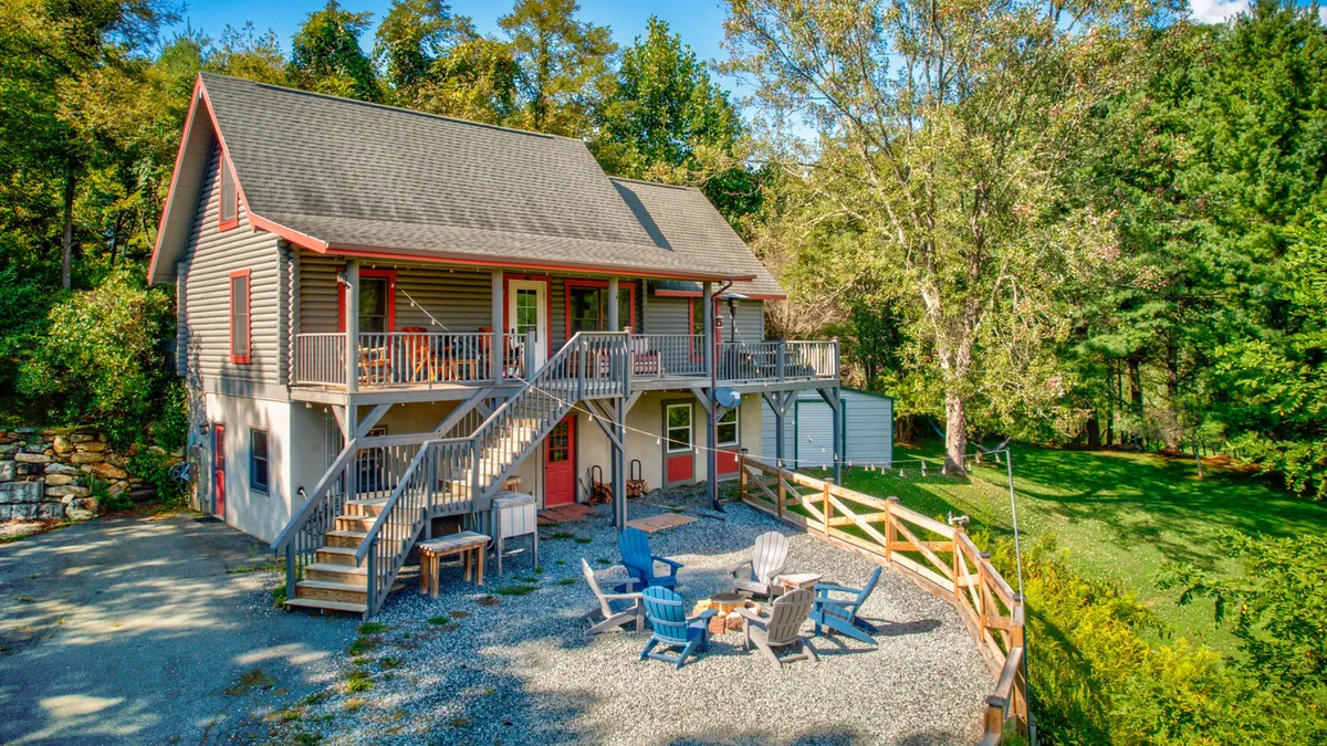 This exterior shot features the lodge as a welcoming two-story house surrounded by lush greenery, with an inviting outdoor seating area and a fire pit at the center. The deck offers expansive views of the natural landscape, creating a sense of adventure and relaxation.