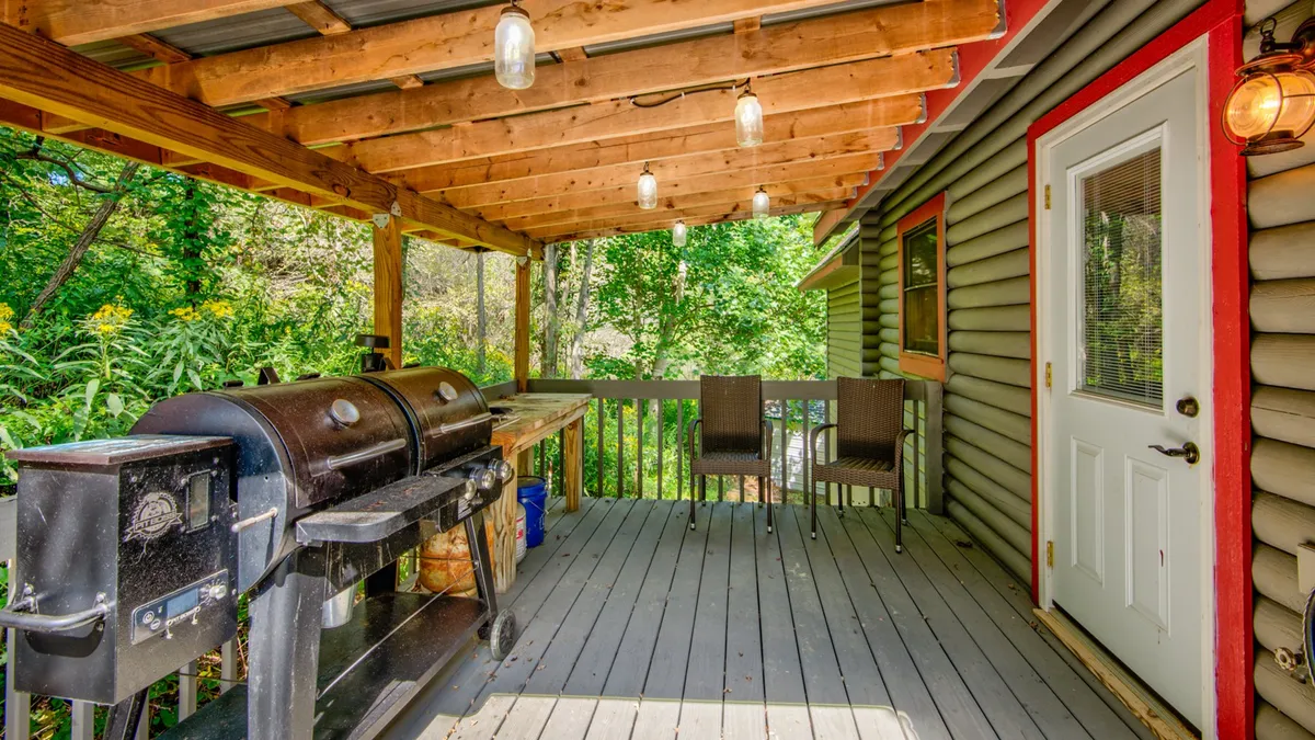 A covered deck featuring a grill and comfortable seating, framed by wooden beams and lush greenery. This inviting outdoor space is ideal for group meals and casual gatherings.