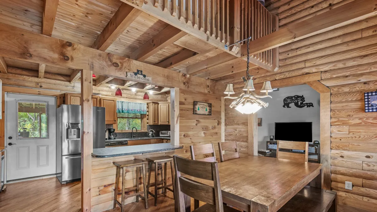 A warm dining area and kitchen are visible, featuring a spacious dining table, a sparkling chandelier overhead, and a welcoming sign near the kitchen. The open layout invites family meals and festive gatherings, with the kitchen seamlessly integrated into the space.