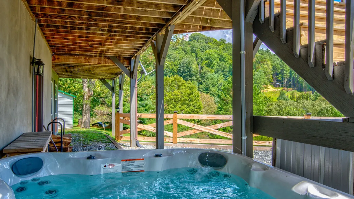 This exterior image showcases the covered patio area with a bubbling hot tub, wooden beams, and lush green foliage surrounding a fenced space. The atmosphere is calm and welcoming, inviting guests to relax in the warmth of the hot tub while enjoying views of nature.