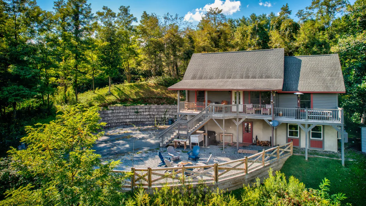 The image showcases the exterior of a cozy, rustic cabin nestled among tall trees. A spacious deck overlooks the woods, and a fire pit set in a gravel area invites guests to gather outdoors. The scene exudes tranquility, with natural surroundings perfect for relaxation or adventure.